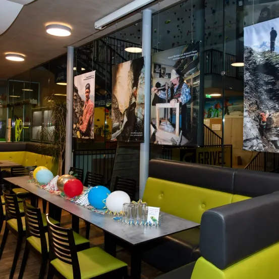 Ein Restaurant mit einem langen Tisch, der mit bunten Luftballons und Gläsern gefüllt ist. Die Wände haben Poster von Menschen und die Decke hat Lampen. Der Boden ist aus Holz.