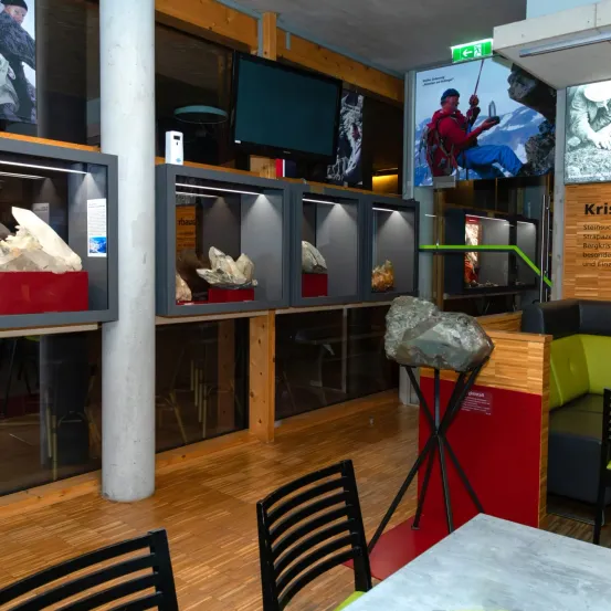 Ein Ausstellungsraum mit Holzböden und Glasvitrinen, der Mineralien präsentiert. Ein großer Stein ist ausgestellt und ein Bildschirm zeigt einen Kletterer.