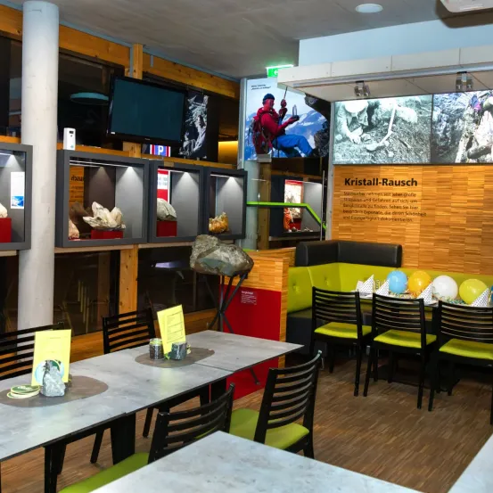 Ein Innenbereich mit einer Essplatzeinrichtung, mit Tischen, Stühlen und Luftballons. Es gibt auch Gesteinsausstellungen und eine Wand mit Text.
