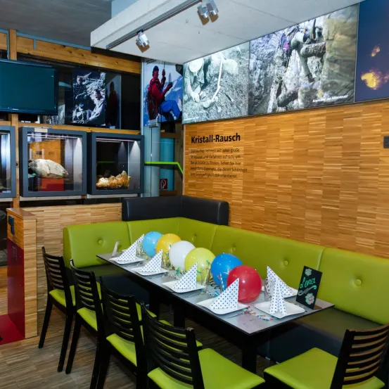 Ein Innenbereich mit einem Tisch voller Luftballons, grünen Polsterstühlen und einer Wand mit einem großen Schild, das Kristall-Rausch sagt. An der Wand befinden sich ein paar Monitore und ein Fernseher, mit Lampen an der Decke.