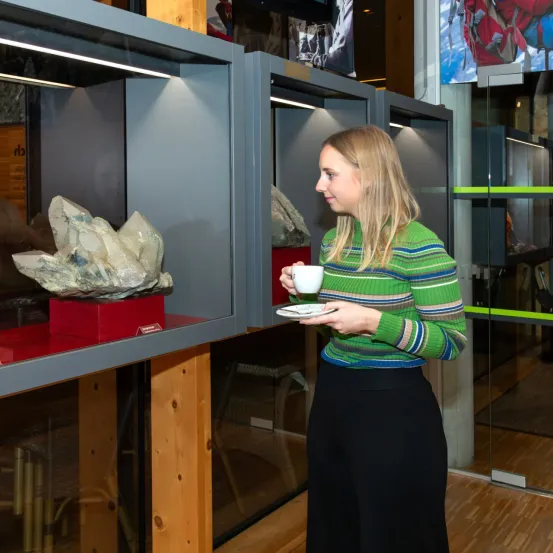 Eine Frau hält eine Tasse mit Untertasse vor einer Glasvitrine mit einem Kristall darin.