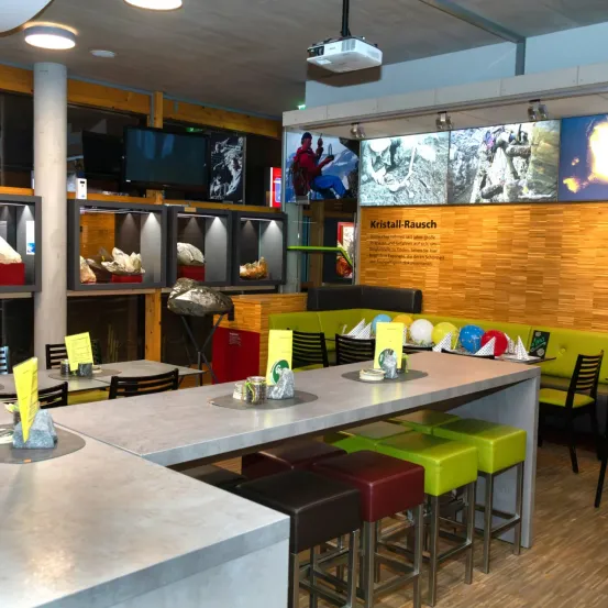 Ein modernes Café mit Theke und Stühlen. Die Wand hat Displays von Kristallen und Monitoren. Die Tische haben gelbe Papiere und verschiedene Gegenstände darauf. Die Decke hat Lampen.