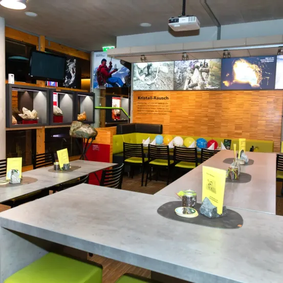 Ein Innenbereich mit einem Betontisch, Stühlen und gelben Luftballons. Eine Projektorlampe befindet sich an der Decke, und ein Schild sagt Kristall-Rausche. Links befindet sich ein Schrank mit Kristallen im Inneren.