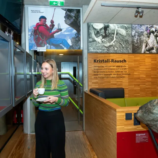 Eine Frau steht in einem Museum, hält eine Tasse und lächelt. Hinter ihr befindet sich eine Wand mit großen Bildern von Kletterern. Die Wand hat ein Schild mit der Aufschrift Kristall-Rausch, mit zusätzlichen Informationen. Es gibt eine Vitrine und einen Felsen auf der rechten Seite.