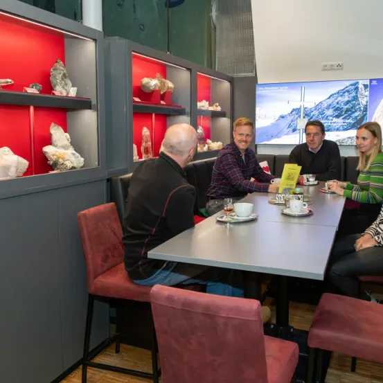 Vier Personen sitzen an einem Tisch mit Tassen und Untertassen, möglicherweise in einem Restaurant. Ein Mann ist links, eine Frau rechts. Der Raum hat einen Holzboden und rot beleuchtete Schränke voller Steine.