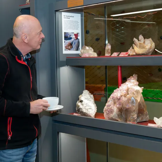 Ein kahlköpfiger Mann in einer schwarzen Jacke hält eine Tasse in der Hand und steht vor einer Glasvitrine mit verschiedenen Kristallen.