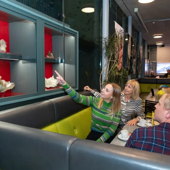 Drei Frauen sitzen in einer Restaurantecke, eine weist auf eine Vitrine mit Skulpturen hin.