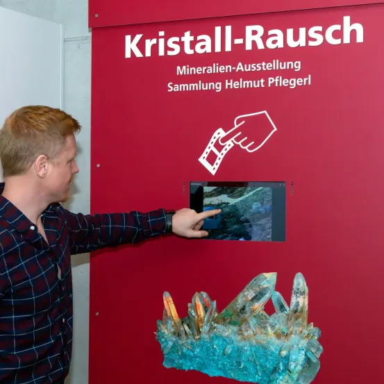 Ein Mann zeigt auf einen Monitor, der ein Bild von Kristallen auf einem roten Schild mit der Aufschrift Kristall-Rausch und Mineralien-Ausstellung Sammlung Helmut Pflegerl anzeigt.