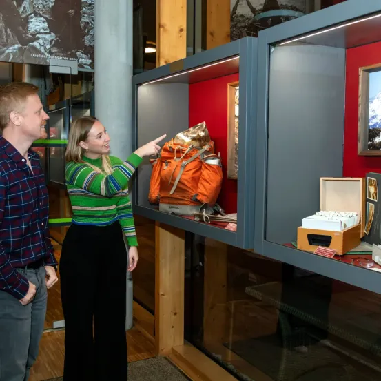 Ein Mann und eine Frau stehen vor einer Vitrine und zeigen auf einen orangefarbenen Rucksack. Der Mann trägt ein kariertes Hemd und die Frau ein grünes Sweatshirt. Dahinter befindet sich ein großes Poster und eine Holzsäule.