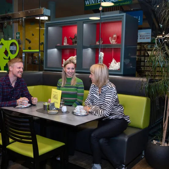 Drei Menschen sitzen an einem Tisch in einem Café. Ein Mann, eine Frau und eine weitere Frau. Dahinter steht eine Pflanze und ein Glasschrank.