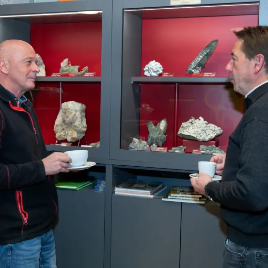 Zwei Männer in einem Raum mit Regalen voller Mineralien, beide halten Tassen und diskutieren möglicherweise über die Exponate.