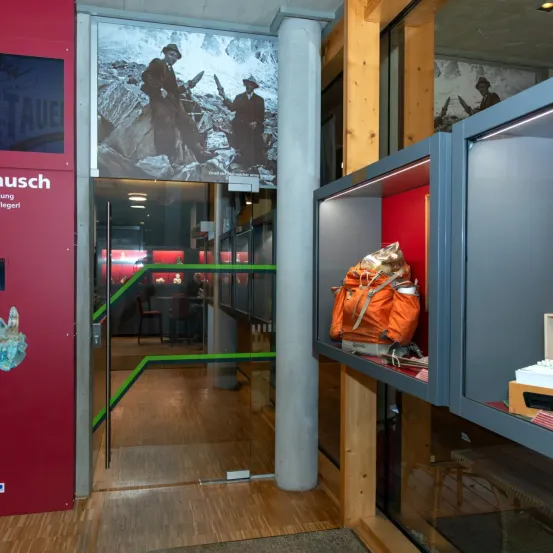 Eine Innenausstellung mit einer Glastür, die ein Foto von zwei Männern in Anzügen und Hüten zeigt, und einem Vitrinenschrank mit einer orangefarbenen Tasche.