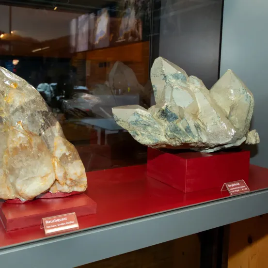 Zwei große Kristalle in einer Glasvitrine ausgestellt. Einer ist mit Rauchtopaz beschriftet und der andere mit Bergkristall. Beide stehen auf roten Sockeln.