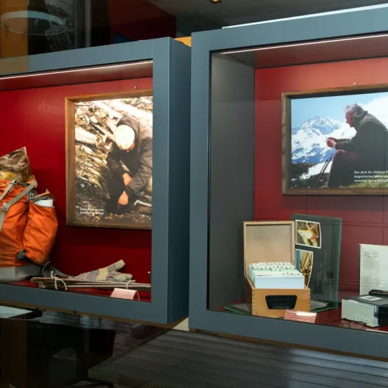 Eine Vitrine mit roter Wand, die ein Foto einer Person in einer verschneiten Landschaft und verschiedene Gegenstände wie einen Rucksack und Bücher zeigt.