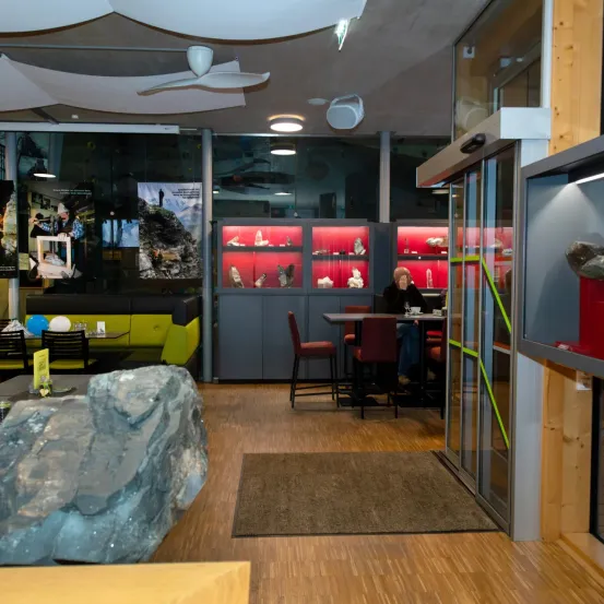 Ein Innenbereich mit Holzböden und -wänden, einem großen Felsen, einem Tisch, Stühlen und einer Vitrine. Eine Person sitzt an einem Tisch. Der Bereich hat Lampen und eine Deckenventilator.