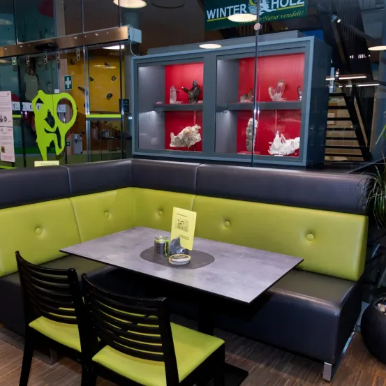 Eine moderne Eckbank mit grünen Kissen und einem grauen Tisch ist zu sehen, zusammen mit einer Glasvitrine mit roten Regalen und verschiedenen Kristallgegenständen.