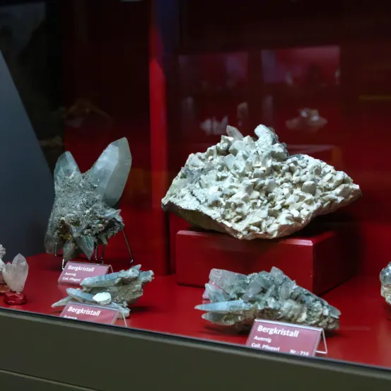 Eine Vitrine mit verschiedenen Kristallen, darunter ein großes Stück mit der Aufschrift Bergkristall, umgeben von kleineren Kristallen. Alle Kristalle stehen auf roten Plattformen.