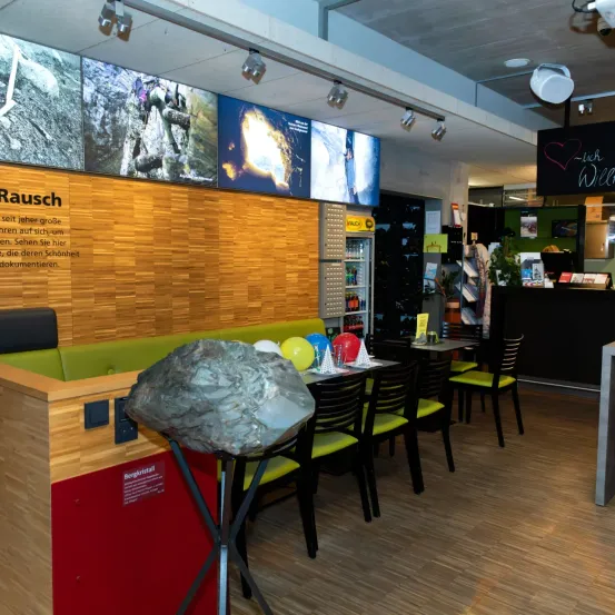 Eine Innenausstellung eines großen grauen Felsens auf einem Ständer, umgeben von Tischen und Stühlen. Es gibt Luftballons und Schilder. Große Bildschirme zeigen Bilder von Felsformationen.