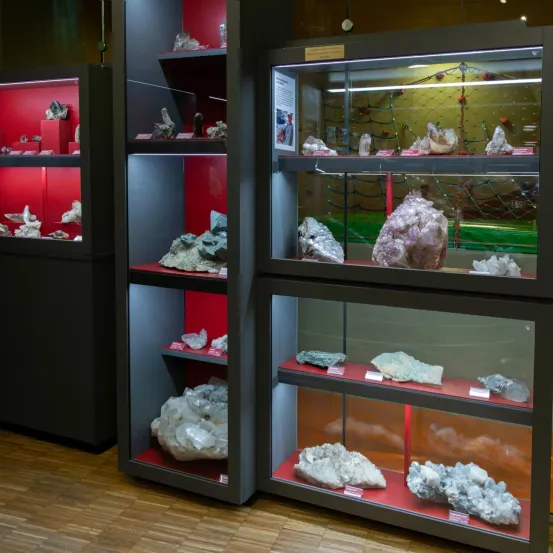 Eine Mineralienausstellung in einem Museum mit drei Glasvitrinen. Die erste Vitrine hat rote Regale, die zweite hat graue und rote Regale und die dritte hat rote Regale mit einigen weißen Etiketten.