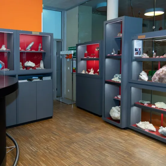 Ein Raum mit Holzboden und mehreren Vitrinen mit Mineralien zur Ausstellung. Die Vitrinen sind grau mit roten Regalen und haben Glasfronten.