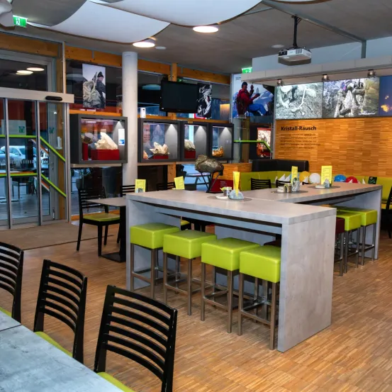 Ein Innenbereich, der einer Bar ähnelt, mit mehreren Stühlen und Hockern um die Tische herum. Eine Projektionsleinwand ist an der Decke montiert. Ein großer Fels ist auf der Seite platziert.