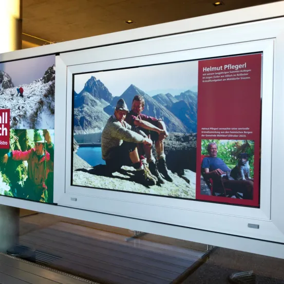 Ein gerahmter Bildschirm zeigt ein Foto von zwei Männern auf einem Berg mit einer Berglandschaft im Hintergrund. Der Bildschirm enthält auch ein Foto eines älteren Mannes, der mit einem Hund sitzt.