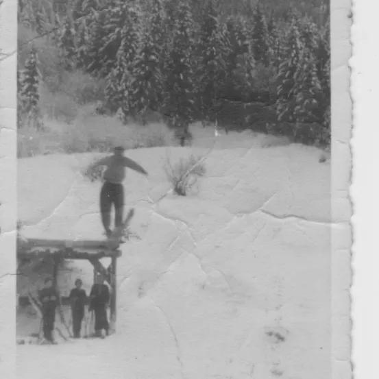 Schwarz-Weiß-Foto einer Person beim Skifahren bergab. Drei Personen stehen in der Nähe einer Holzstruktur am Fuße des Berges.