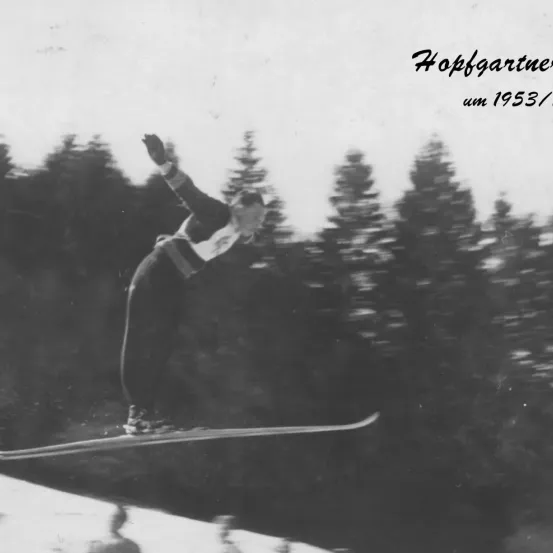 Ein Schwarz-Weiß-Bild eines Skifahrers in der Luft, datiert um 1953. Der Skifahrer trägt einen Helm und Skiausrüstung.