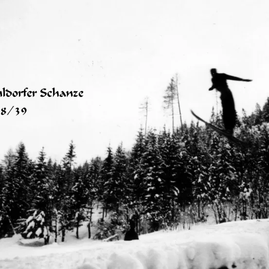 Ein Schwarz-Weiß-Foto eines Skifahrers, der von einem schneebedeckten Hang namens 'Indorfer Schanze 8/39' springt. Umgeben von schneebedeckten Bäumen.