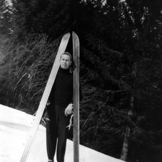 Ein Schwarz-Weiß-Bild eines Mannes, der auf einem verschneiten Hang mit Skiern steht. Er ist in einem Rollkragenpullover und Hose gekleidet.
