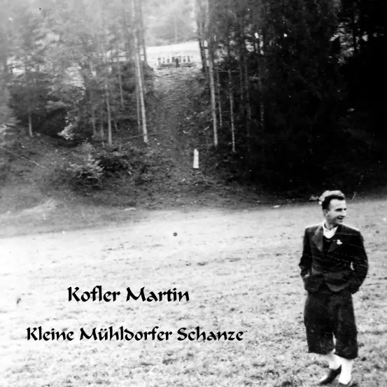 Ein Schwarz-Weiß-Bild zeigt einen Mann namens Martin Koffler, der in einem Grasfeld steht. Hinter ihm befinden sich Bäume und ein kleiner Hügel mit einem Pfad.