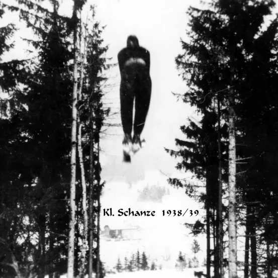 Ein Skispringer wird 1938/39 in der Luft auf der K1. Schanze Skisprungschanze umgeben von hohen Bäumen und Schnee erfasst.