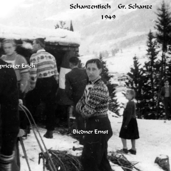 Ein Schwarz-Weiß-Foto aus dem Jahr 1949 zeigt Menschen in einer verschneiten Landschaft. Im Vordergrund steht eine Frau namens Ernst. Dahinter versammeln sich mehrere Personen in der Nähe einer Holzstruktur. Kiefern umgeben das Gebiet, und der Himmel ist bedeckt.