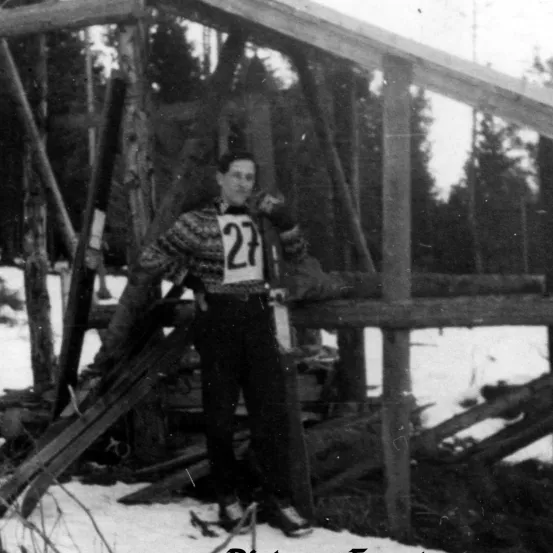 Ein altes Schwarz-Weiß-Foto zeigt einen Mann im Schnee mit einem Skisprung im Hintergrund. Er trägt einen Pullover mit der Nummer 27.