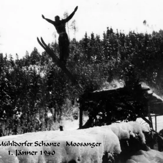 Ein Skispringer ist in der Luft in einer verschneiten Landschaft mit Kiefern, über der Mühldorfer Schanze Moosanger Sprungschanze, datiert auf den 1. Januar 1940.