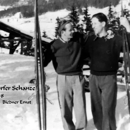 Zwei Männer stehen auf einem verschneiten Berg mit Kiefern im Hintergrund. Ein Mann trägt einen Hut, und beide halten Skistöcke.