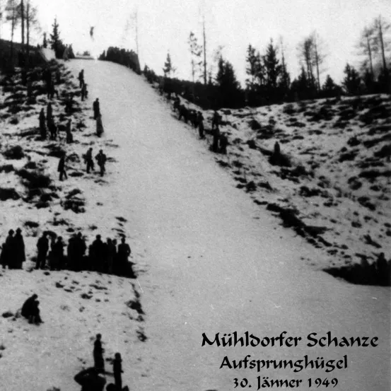 Ein Schwarz-Weiß-Bild zeigt Menschen, die auf einem verschneiten Hang stehen. Einige sind unten versammelt, während andere entlang des Hangs verstreut sind. Bäume sind im Hintergrund zu sehen. Das Bild hat eine Überlagerung mit Text auf Deutsch.