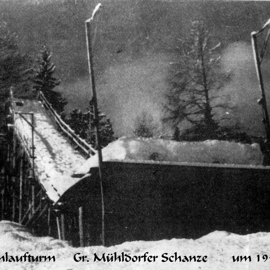 Ein Schwarz-Weiß-Foto einer Skisprungschanze, möglicherweise die Gr. Mühldorfer Schanze, um 1930. Bäume und eine verschneite Landschaft sind sichtbar.