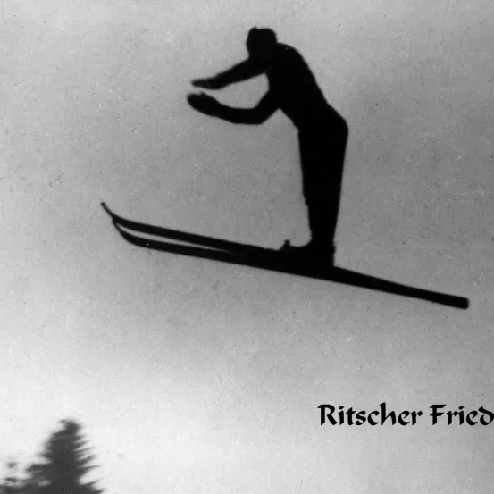 Ein Schwarz-Weiß-Foto eines Mannes in der Luft beim Skifahren. Er trägt einen Hut und hält Skistöcke. Unten steht 'Ritscher Fried'.