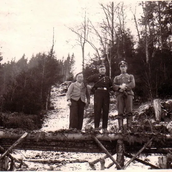 Drei Personen stehen auf einer Holzbrücke in einem verschneiten Wald. Einer trägt eine militärische Uniform, die anderen zivile Kleidung.