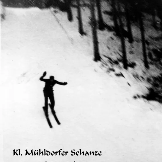 Ein Skispringer in der Luft auf der Muhldorfer Schanze. Der schneebedeckte Hügel und die umliegenden Bäume sind sichtbar.