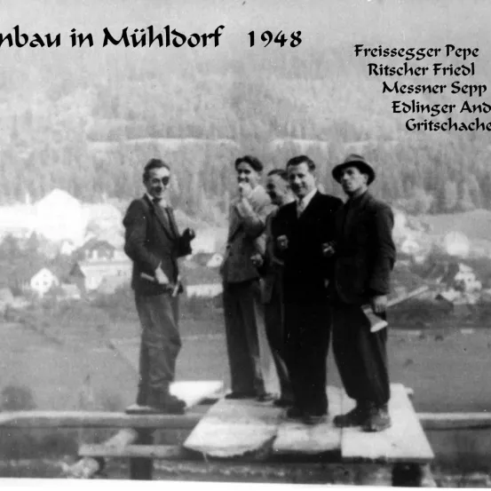 Ein Schwarz-Weiß-Foto von 1948 zeigt vier Männer, die auf einer Holzplattform stehen, die als Freissegger Pepe, Ritscher Friedl, Messner Sepp und Edlinger And Gritschacher beschriftet ist. Sie befinden sich in Mühldorf.