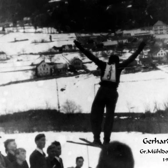 Ein Schwarz-Weiß-Foto eines Skifahrers inmitten eines Sprungs in den verschneiten Bergen. Der Skifahrer trägt die Nummer '1' auf der Brust. Unten beobachten mehrere Zuschauer. Das Foto ist auf das Jahr 1919 datiert.