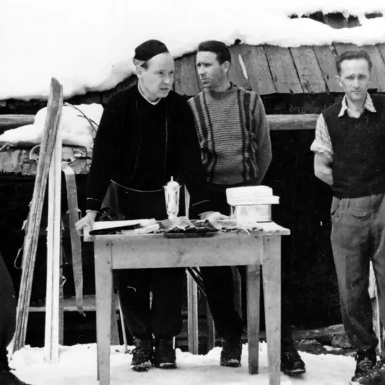 Ein Schwarz-Weiß-Foto von drei Männern, die in einem verschneiten Bereich in der Nähe eines Tisches stehen, mit einer Holzstruktur und Skistöcken im Hintergrund.