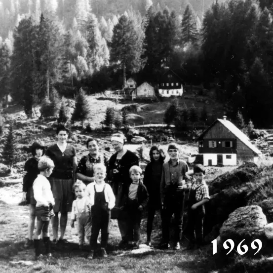 Ein Schwarz-Weiß-Foto aus dem Jahr 1969 zeigt eine Gruppe von Kindern und Erwachsenen vor einer bergigen Waldkulisse. Das Jahr ist unten rechts angezeigt.