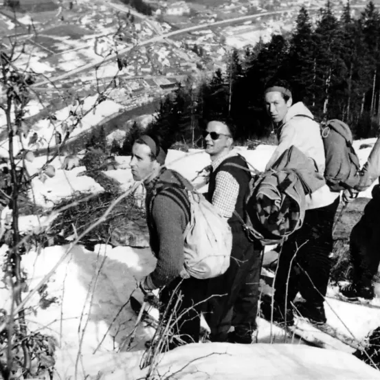 Ein Schwarz-Weiß-Foto von vier Männern, die in schneebedecktem Gelände wandern, Winterkleidung tragen und Rucksäcke tragen.