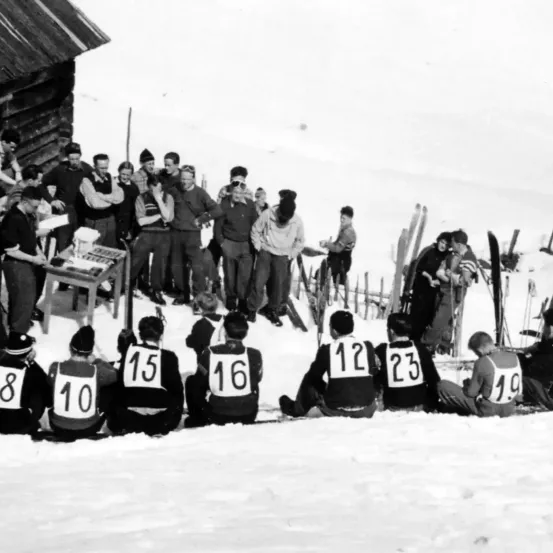 Ein Schwarz-Weiß-Foto zeigt eine Gruppe von Menschen in Winterkleidung, einige stehend und andere sitzend im Schnee. Sie tragen nummerierte Westen, wobei die Nummern 12, 15, 16, 19, 10 und 8 sichtbar sind. Dahinter steht ein kleines Gebäude.