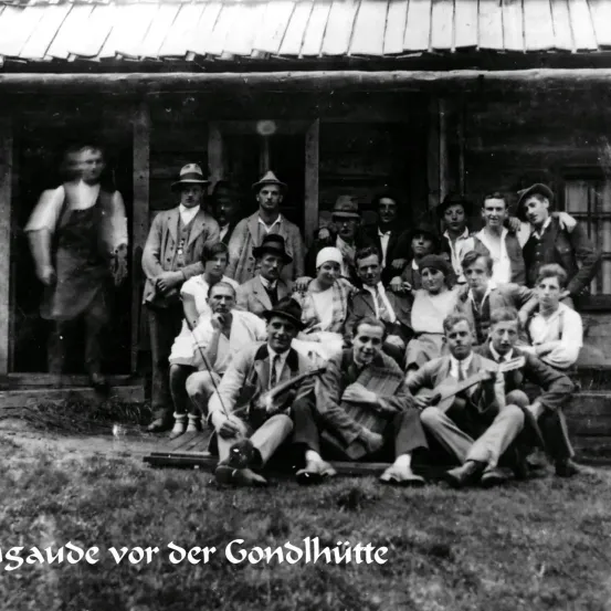 Eine Gruppe von Menschen steht und sitzt vor einem Holzhaus. Sie sind in altertümlicher Kleidung gekleidet und scheinen für ein Foto zu posieren. Der Text unten lautet 'saunde vor der Gondelhutte.'
