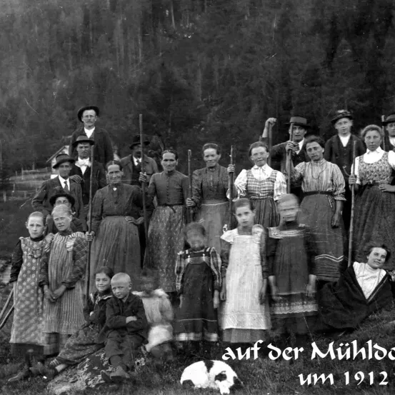 Ein Schwarz-Weiß-Foto zeigt eine Gruppe von Menschen aus der Vergangenheit, die im Freien stehen und sitzen. Einige halten Stöcke, und ein Hund ist anwesend. Das Bild ist mit 'um 1912' beschriftet.