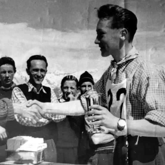 Ein Schwarz-Weiß-Foto von einer Gruppe von Männern in Skiausrüstung, einer von ihnen hält einen Pokal in der Hand und schüttelt einem anderen die Hand.